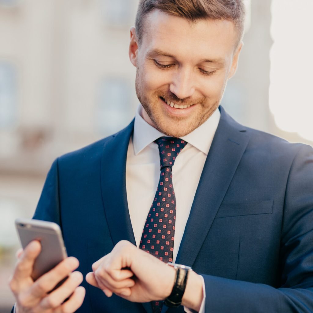Photo of clever male lawyer waits for client outdoor, looks at wristwatch, holds smart phone as waits for important call, wears formal suit, has pleasant smile on face. People, time, meeting concept
